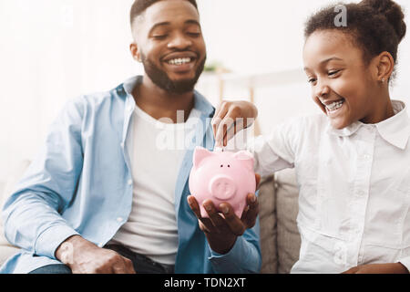 Risparmi personali. Piccola ragazza afro mettere il denaro a Piggybank Foto Stock