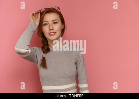Redhead ragazza con treccia rosso sorrisi a fotocamera mettendo occhiali sul suo capo con una mano in posa su sfondo rosa Foto Stock