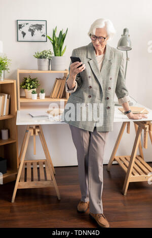 Elegante bianco ritirato dai capelli donna in abito grigio texting nello smartphone mentre in piedi da scrivania con forniture di lavoro Foto Stock