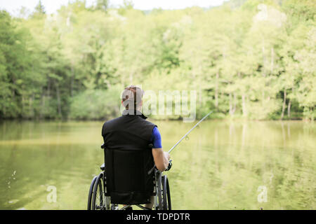 Felice e giovani disabili o handicappati uomo pesca in un lago in natura. Sport popolari per paraplegici. Foto Stock