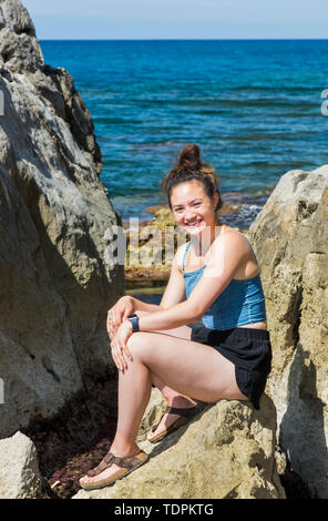 Un giovane turista femminile siede su rocce lungo la costa mediterranea; Cefalu, Sicilia, Italia Foto Stock