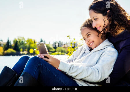 Una mamma e sua figlia prendendo un autoritratto e controllare i social media mentre si riposa accanto a un lago in un parco della città in una calda giornata di caduta Foto Stock