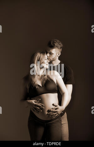 Una giovane coppia in attesa di guardare tutti gli altri e che il padre è la bacia sulla fronte e tenendo premuto il suo ventre in uno studio su uno sfondo scuro Foto Stock