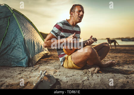 Felice giovane musicista ha un grande tempo in spiaggia. Egli è seduto sul fiume, riproduzione di ukulele e cantare al tramonto. Foto Stock