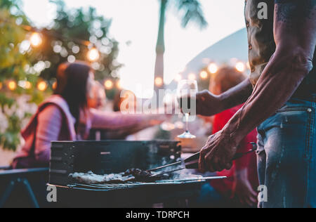 La famiglia felice di fare cena a barbecue sul tetto di notte - vicino la mano di uomo tatuaggio per la cottura alla griglia per il barbecue all'aperto Foto Stock