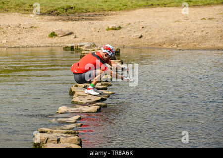 OGMORE DAL MARE, GALLES - Aprile 2019: Persona in abbigliamento ciclo di scattare una foto su un telefono cellulare mentre in equilibrio su le pietre miliari attraverso il fiume Foto Stock