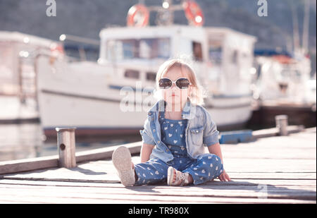 Elegante ragazza ragazzo indossa alla moda abbigliamento denim seduta sul mare in legno pier all'esterno. Guardando alla fotocamera. Infanzia. Foto Stock