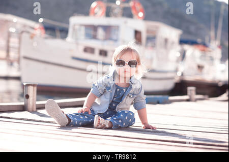 Eleganti bambina indossa abbigliamento denim seduta sul molo sul mare all'esterno. Guardando alla fotocamera. Infanzia. Foto Stock