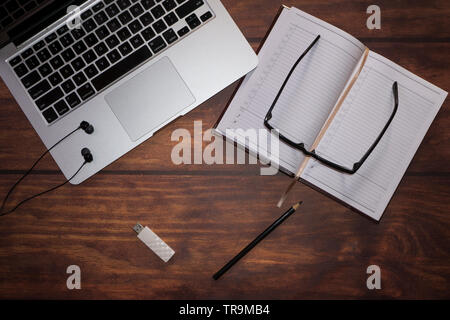 Lay piatto laptop, notebook, penne, unità flash, i bicchieri e le cuffie sul marrone texture di legno sullo sfondo Foto Stock