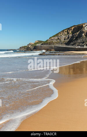 Un'onda rotoli attraverso la sabbia all'estremità meridionale della spiaggia di Newcastle. La passeggiata costiera nella distanza conduce al King Edward Park e il bar sulla spiaggia. Newcast Foto Stock