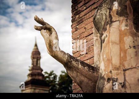 Statua di Buddha in Sukhothai parco storico con le dita in spirituale mudra gesto apparentemente toccando la guglia di un antico stupa in background Foto Stock