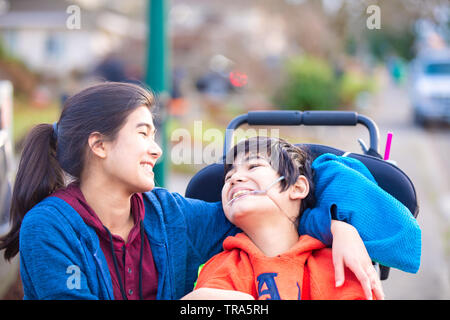 Biracial big sister amorevolmente abbracciando disabilitato piccolo fratello in wheelchairoutdoors, sorridente Foto Stock