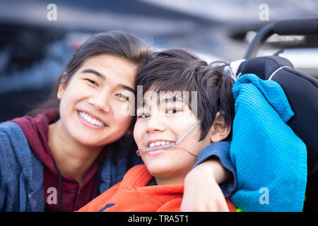 Biracial big sister amorevolmente abbracciando disabilitato piccolo fratello in wheelchairoutdoors, sorridente Foto Stock