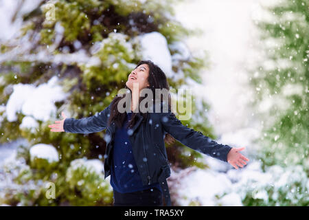 Biracial teen ragazza vestita di nero giacca di pelle all'esterno in inverno tempesta di neve, le braccia aperte e il sollevamento a faccia in su verso il cielo, godendo di fiocchi di neve f Foto Stock