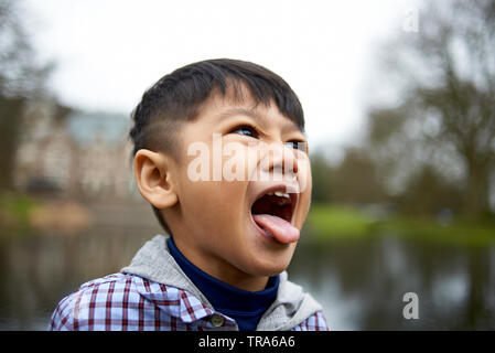 Ritratto di un adorabile piccolo ragazzo asiatico con grandi occhi marroni in un parco fuori la sua lingua nella frustrazione