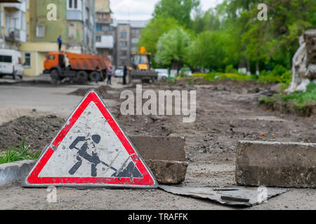 Segno di riparazione su strada si erge sullo sfondo del distrutto asfalto nel cortile di un edificio a più piani, attrezzature da costruzione e wo Foto Stock