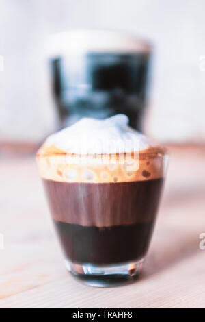 Doppio macchiato, espresso con schiuma di latte in un bicchiere piccolo di fronte ad una pinta di stout con una schiuma sulla parte superiore in background su un tavolo di legno. Foto Stock