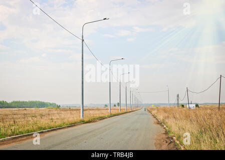 Strada asfaltata e lanterne sul lato della strada e un ciclista solitario nella distanza nel caldo sole. Il concetto di viaggi e avventura. Foto Stock