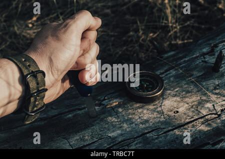 Man mano sporca detiene un coltello, una bussola su un vecchio log in foresta.La sopravvivenza. Foto Stock
