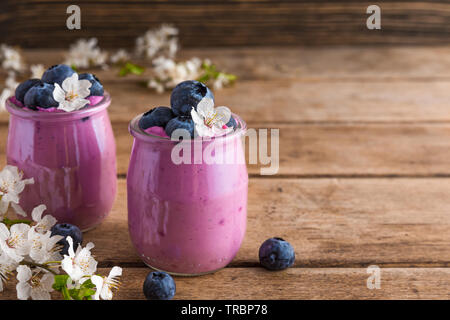 Una sana prima colazione. Mirtillo yogurt in bicchieri serviti con mirtilli freschi e la primavera sbocciano i fiori ciliegio fiori su tavola in legno rustico. close up Foto Stock