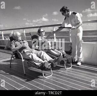 1960s, storico, un cameriere maschile in uniforme serve bevande a due passeggeri adulti che si rilassano in sedie reclinabili all'esterno sul ponte della nave da crociera/nave da crociera norvegese dell'oceano, Sagafjord. Foto Stock