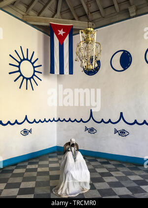 Afro-caraibica religione bambola vestito con i tradizionali abiti bianchi come seguaci della religione Santeria carrier seduto sotto una bandiera cubana in una casa Foto Stock