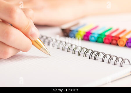 Mano d'uomo con un nero-penna d'oro su un foglio pulito di notebook a scacchi. Sullo sfondo sono colorati pennarelli. Foto Stock