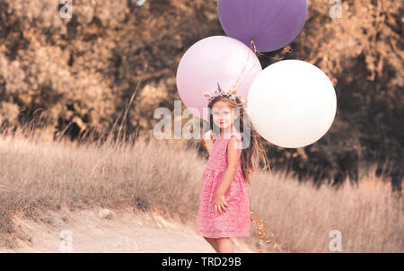 Sorridente ragazza di capretto 4-5 anni palloncini di contenimento in campo. Indossando estate elegante abito rosa e archetto unicorn all'esterno. Guardando alla fotocamera. Infanzia Foto Stock