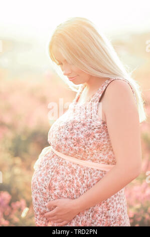 Donna incinta in piedi nella luce solare all'esterno. Tenendo la pancia. indossare abiti eleganti con motivo floreale. La maternità. La maternità. Foto Stock