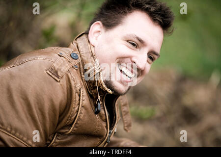 Ritratto di un giovane simpatico uomo vestito con una giacca di pelle seduta su una panchina nel parco Foto Stock