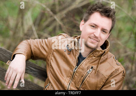 Ritratto di un giovane simpatico uomo vestito con una giacca di pelle seduta su una panchina nel parco Foto Stock