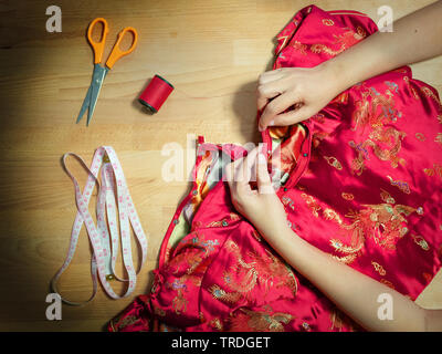 Vista dall'alto ritagliata di mani femminili cucito Cheongsam abito con ago a sarta workplace con attrezzatura per cucire Foto Stock