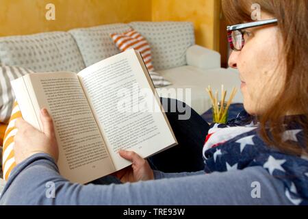 Donna di mezza età con i capelli rossi con gli occhiali seduto su un divano nella lettura di un libro Foto Stock