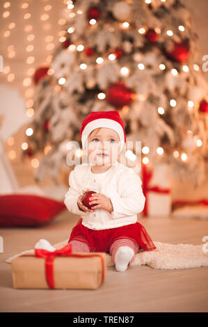 Sorridente bambina 1 anno vecchio seduto sul pavimento con le decorazioni di Natale su luci in background. Stagione di vacanze. Infanzia. Foto Stock