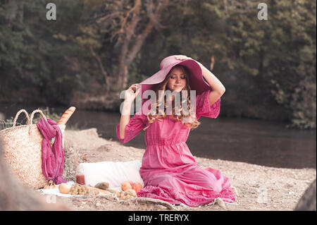 Elegante ragazza adolescente indossando abiti estivi e grande cappello avente un picnic con frutti all'esterno. Sorridente ragazza seduta nella foresta. Foto Stock