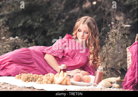 Ragazza bionda giacente avente picnic all'aperto. Indossando elegante abito rosa. Foto Stock