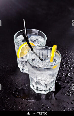 Due bicchieri di freddo fresca acqua gassata con cubetti di ghiaccio e le fette di limone. Acqua di soda sul bagnato su sfondo nero. Foto Stock