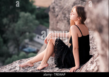 Blonde teen ragazza 14-16 anno indossando abito nero in appoggio all'esterno. Foto Stock