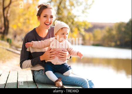 Felice azienda madre bambina di 1 anno indossando maglioni a maglia seduta sul molo di legno in autunno park. Guardando lontano. La felicità. Foto Stock