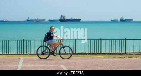 Femmina ciclista ricreative in sella alla sua moto lungo la East Coast Park, navi da carico in background di Singapore. Foto Stock