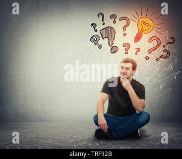 Malinconici uomo seduto sul pavimento tenendo la mano sotto il mento ha molte domande come segni di interrogazione al di sopra della testa e una brillante idea come lampadina incandescente Foto Stock