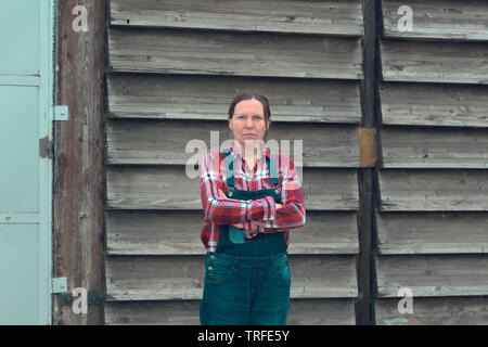 Ritratto di donna agricoltore di fronte al casale capannone. Donna vestita di plid camicia e jeans denim tuta come salariato agricolo con le braccia incrociate Foto Stock