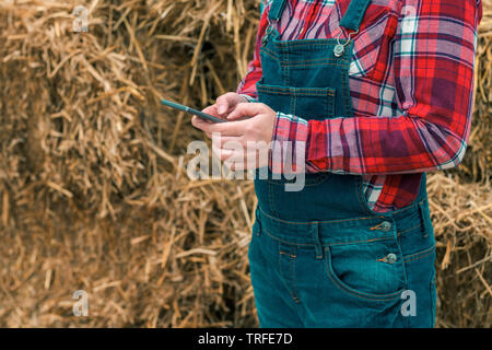 L'agricoltore femmina la digitazione di un messaggio sms sul cellulare di fronte al casale pagliaio, close up delle mani Foto Stock
