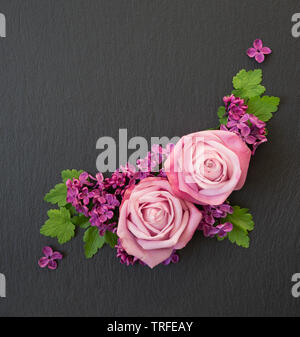 Composizione decorativa consistente di rose rosa, lilla viola fiori e foglie verdi su un nero ardesia con copy-spazio. Appartamento laico, vista dall'alto, overhead Foto Stock