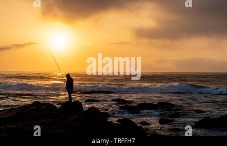 Pescatore solitario sulle rocce durante la bellissima alba sulla spiaggia. Durban, Costa Orientale, Sud Africa Foto Stock