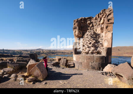 Giovane donna che guarda ad una delle imponenti torri di sepoltura chiamato chullpas di Sillustani con il lago Umayo in background, Regione di Puno, Perù Foto Stock