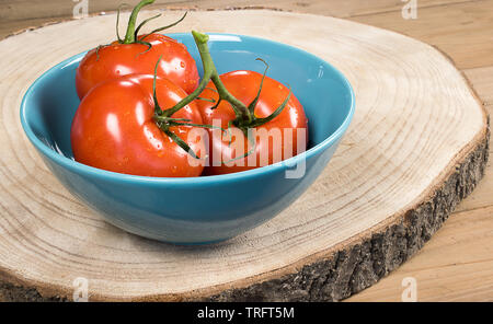 Un gruppo di tre freschi pomodori interi in una ciotola blu su una tavola di legno tronco di albero decorato con foglie di basilico. Foto Stock