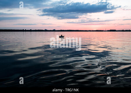 Un pescatore solitario in una barca sul lago al tramonto Foto Stock