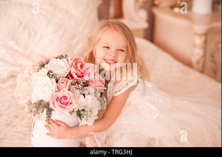 Laughing baby ragazza con fiori seduta nel letto. Indossando elegante abito bianco. Guardando lontano. Festa di compleanno. Foto Stock