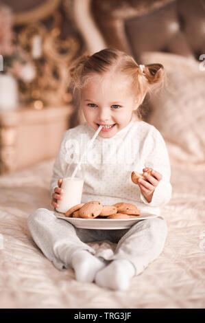 Laughing baby girl 3-4 anni latte drking mangiare i cookie nel letto vicino. Buona mattina. Infanzia. Foto Stock